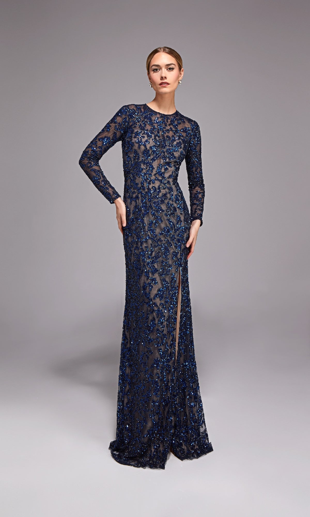 A woman poses against a plain grey background wearing the Alyce 27788 Long Formal Dress in Navy/Sand—a beaded, floor-length gown with long sleeves and a high slit.
